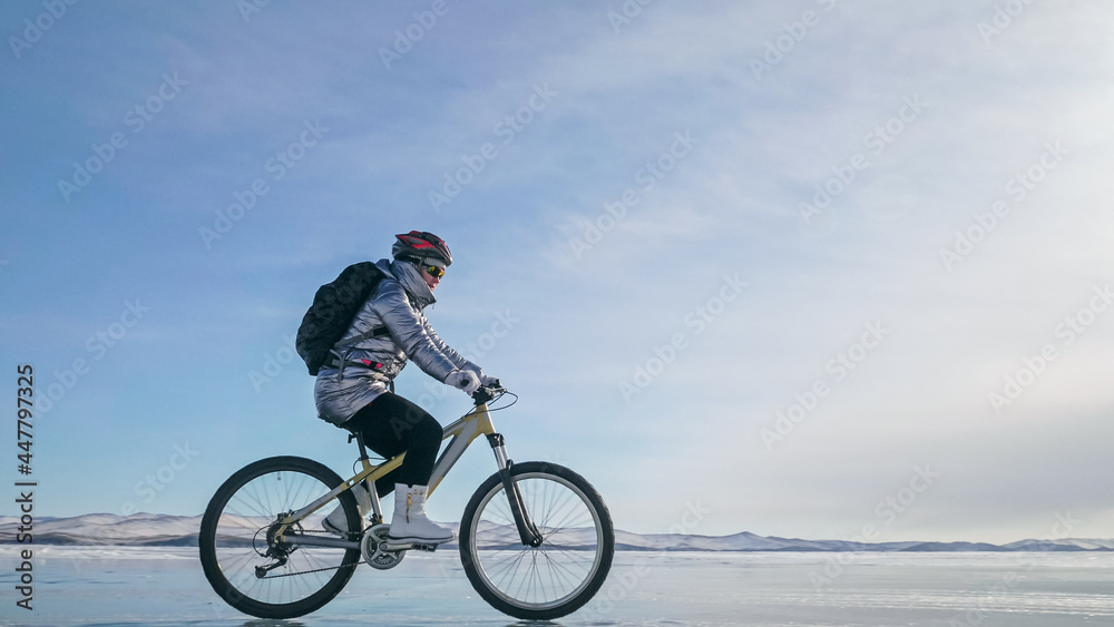 Woman is riding bicycle on the ice. Girl is dressed in a silvery down jacket, cycling backpack and helmet. Ice of the frozen Lake Baikal. Tires on bike are covered with spikes. Traveler is ride cycle. Woman is riding bicycle on the ice. Girl is dressed in a silvery down jacket, cycling backpack and helmet. Ice of the frozen Lake Baikal. Tires on bike are covered with spikes. Traveler is ride cycle.