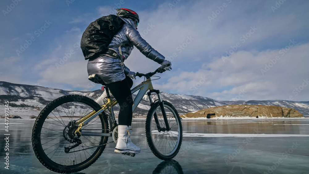 Obraz premium Woman is riding bicycle on the ice. Girl is dressed in a silvery down jacket, cycling backpack and helmet. Ice of the frozen Lake Baikal. Tires on bike are covered with spikes. Traveler is ride cycle.