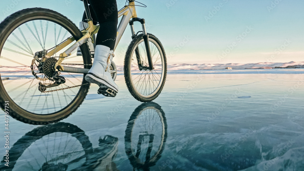 Fototapeta premium Woman is riding bicycle on the ice. Girl is dressed in a silvery down jacket, cycling backpack and helmet. Ice of the frozen Lake Baikal. Tires on bike are covered with spikes. Traveler is ride cycle.