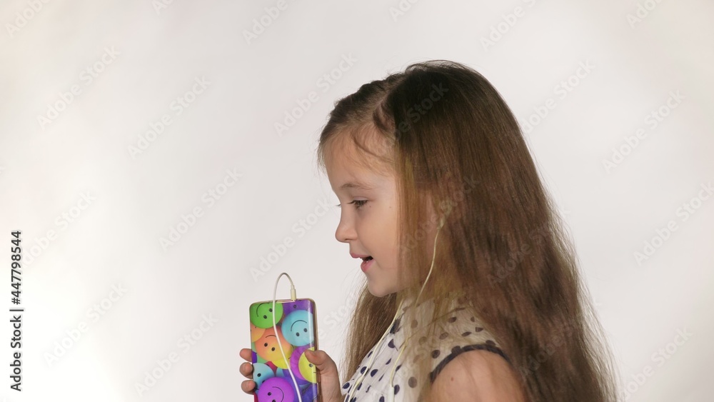 Child with earflaps dancing at studio background. The girl listens to music on the smartphone. The kid has loose long hair. Beautiful cute happy little girl listens to music on Headphones. Child with earflaps dancing at studio background. The girl listens to music on the smartphone. The kid has loose long hair. Beautiful cute happy little girl listens to music on Headphones.