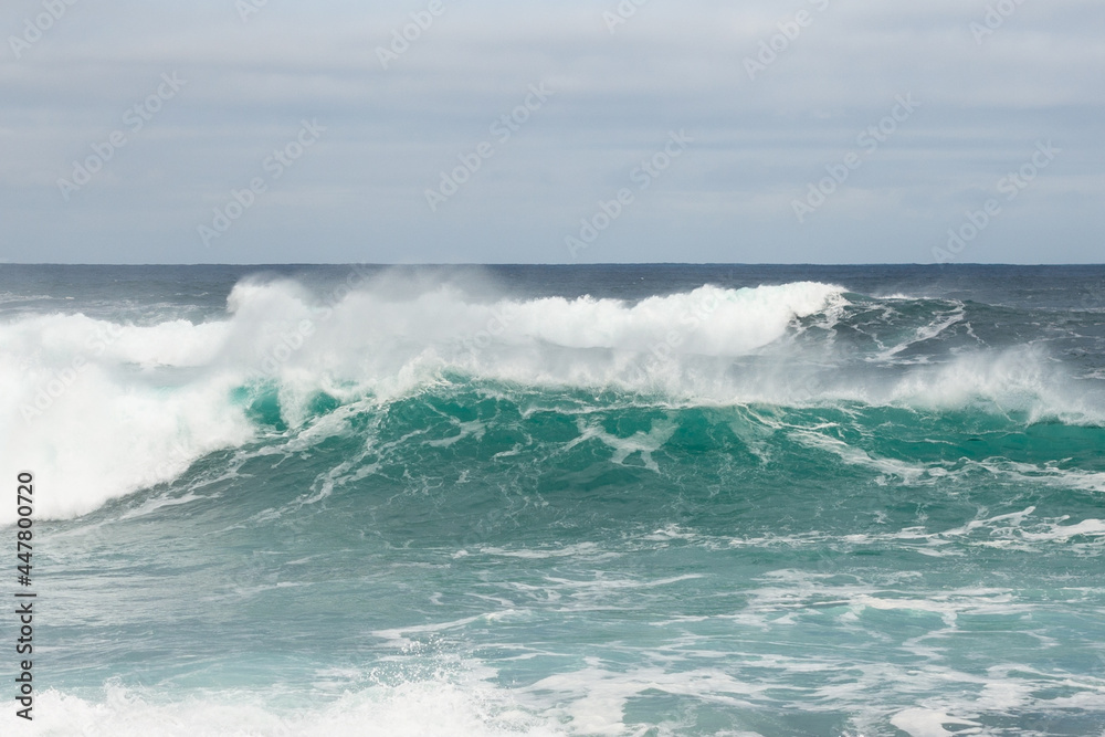 An angry turquoise green color massive rip curl of a wave as it rolls along a beach. The white mist and froth from the wave are foamy and fluffy. The Atlantic Ocean in the background is deep blue. 