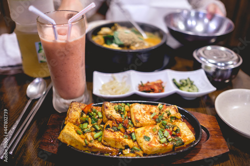 Delicious traditional korean meal with a pan with grilled tofu and vegetables, "banchan" (small side dishes) and a strawberries smoothie, and a girl with a plate of Kimchi stew ("Kimchi-jjjigae")