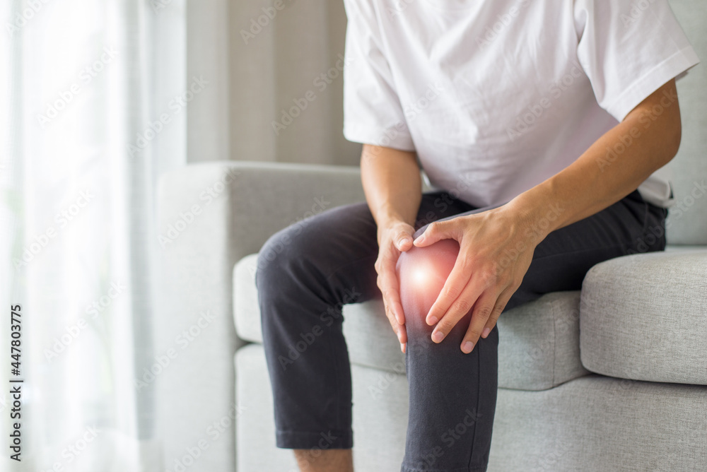 Obraz premium A young man wearing a white T-shirt and gray pants sits on the sofa in the living room at home. He felt pain in his knee area, so he used both hands to hold his knee.