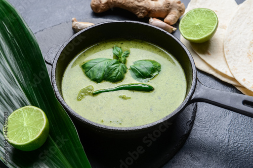 Pot with tasty green curry and ingredients on dark background