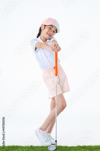 Portrait isolated studio shot of pretty cute little Asian golfer in sport uniform and colorful cap stand on green grass posing put hands on orange grip of golf club driver in front white background
