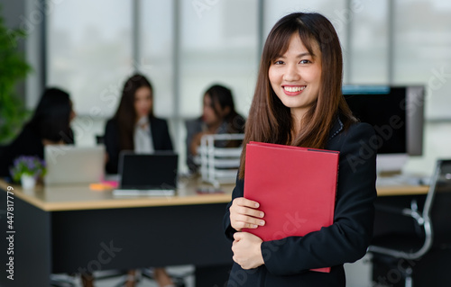 Portrait of young attractive Asian female office worker in formal business suits  smiling at camera in office with blurry colleagues sitting in office as background