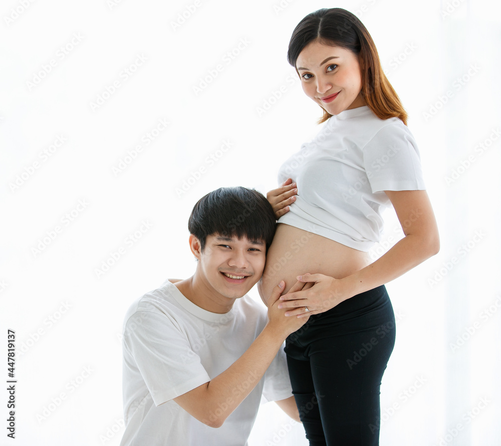 young lovely Asian husband and wife stay together in the living room. A pregnant woman standing and looking at her belly while a man sitting and listening to a baby in her belly with love copy space young lovely Asian husband and wife stay together in the living room. A pregnant woman standing and looking at her belly while a man sitting and listening to a baby in her belly with love copy space