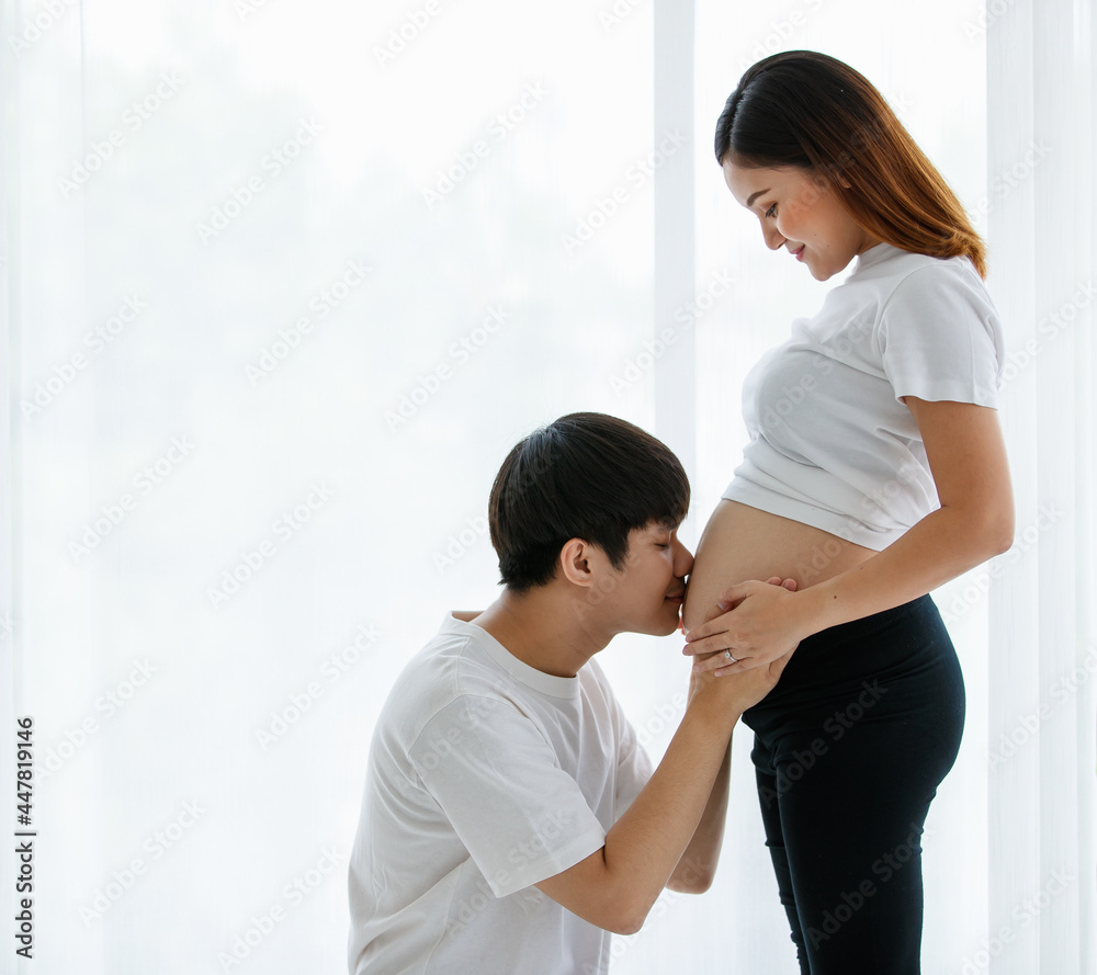portrait of a very lovely young Asian couple. A husband sitting and kissing a wife's belly while a pregnant woman standing smiling and looking at a man with much love portrait of a very lovely young Asian couple. A husband sitting and kissing a wife's belly while a pregnant woman standing smiling and looking at a man with much love