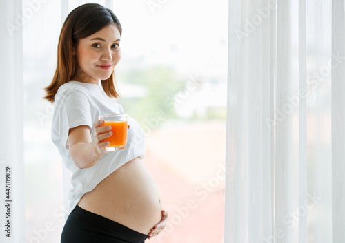 Free Download portrait of a good-looking Asian pregnant woman dress casually standing and touching her belly with love and happiness while looking and smiling at a camera - GFXLoad