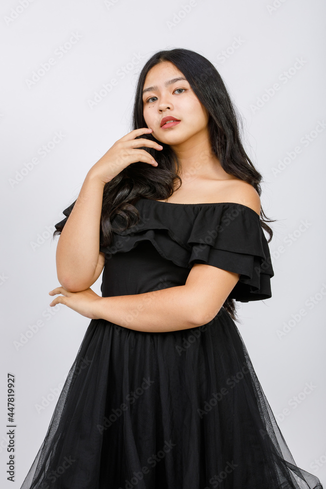 Vertical portrait shot of a cute smiling young girl teenager in a black dress looking at camera in studio isolated white background. Junior girl in fashionable clothes touching hair with shy emotion Vertical portrait shot of a cute smiling young girl teenager in a black dress looking at camera in studio isolated white background. Junior girl in fashionable clothes touching hair with shy emotion