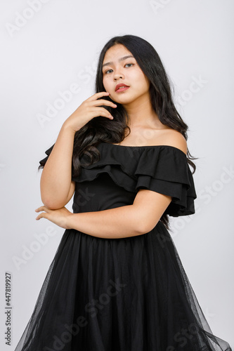 Vertical portrait shot of a cute smiling young girl teenager in a black dress looking at camera in studio isolated white background. Junior girl in fashionable clothes touching hair with shy emotion