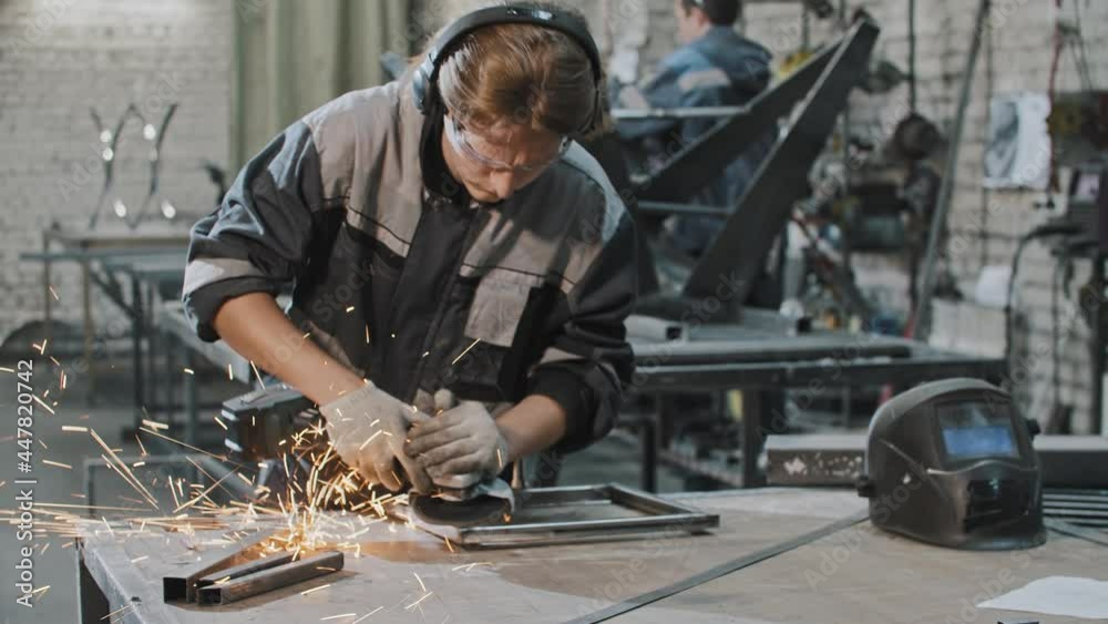 Young man in protective glasses working at the plant - grinding the metal construction with angle grinder Young man in protective glasses working at the plant - grinding the metal construction with angle grinder