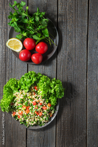 There are two dark plates on a dark wooden background. In one, cooked tabouleh salad, in another plate, tomatoes, lemon and mint branches. View from above.Copy Space.