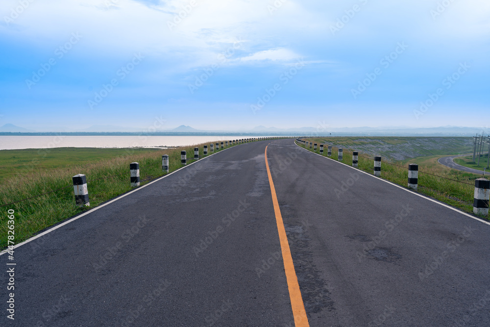 pavement asphalt road view leads to the horizon at Pasak Chonlasit Dam in Lopburi, Thailand. beautiful scenery of road with grass field and water lake