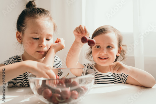Happy children eat summer berries. Delicious bright juicy berries. Children in striped T-shirts eat. Two sisters eat cherries for an afternoon snack. Children sit at the table and eat