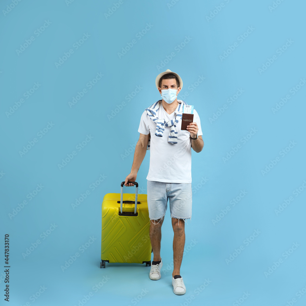 Male tourist in protective mask holding passport with ticket and suitcase on turquoise background Male tourist in protective mask holding passport with ticket and suitcase on turquoise background