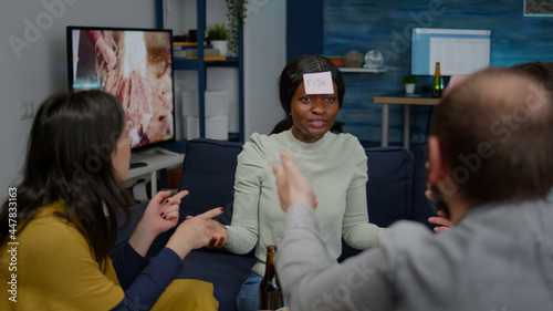 Multiethnic friends having attaching sticky notes on forehead while playing guess who game. Mixed race people having fun, laughing together while sitting on sofa in living room late at night