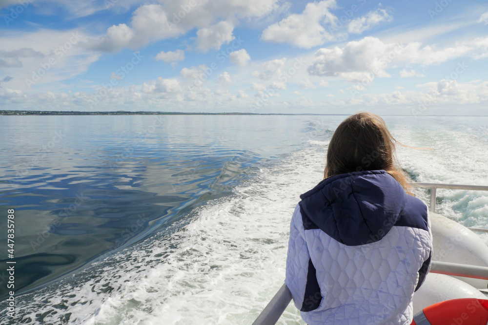 Fototapeta premium Teenager girl on a ocean voyage watching ship trail and ocean scenery. Galway bay, Ireland. Travel and tourism concept. Blue cloudy sky. The model in white jacket, has long hair