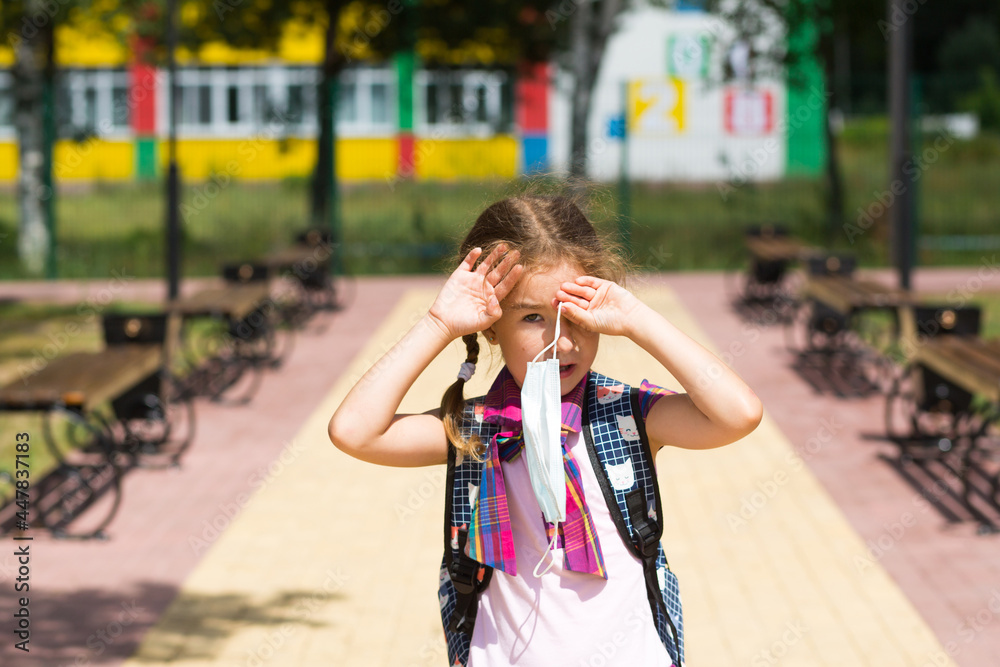 Fototapeta premium Girl with a backpack near the school after classes takes off a medical mask, unhappy, tired and headache. He holds his head with his hands, there is a lack of oxygen. Coronavirus, security measures