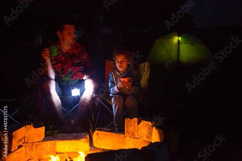 Dad and daughter sit at night by the fire in the open air in the summer in nature. Family camping trip, gatherings around the campfire. Father's Day, barbecue. Camping lantern and tent