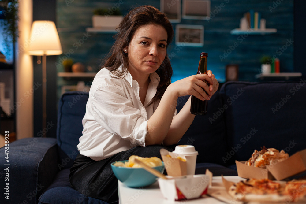 Portrait of cheerful woman sitting on couch smiling at camera while enjoying home-delivered late at night in living room. Caucasian female holding beer bottle. Takeaway food delivery
