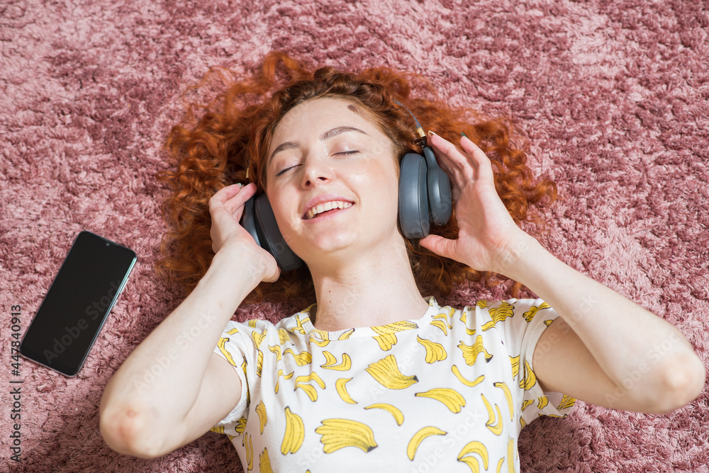 Beautiful caucasian girl in headphones lie on the floor on the carpet at home and listening to music