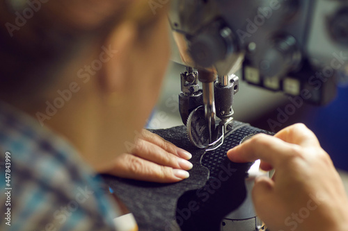 Female worker making detail for new footwear on industrial sewing machine, needle and hands holding piece of textile in closeup. Manufacturing industry and production process at shoe factory concept