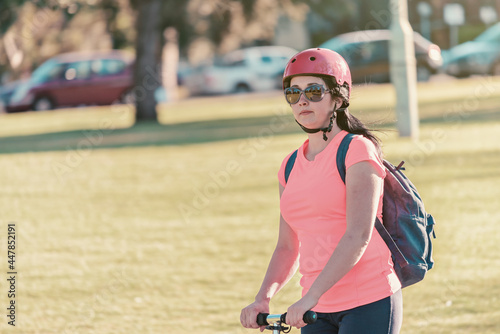 Portrait of a young woman riding push scooter through Adelaide Parklands while  wearing sunglasses with a pink top and bike helmet