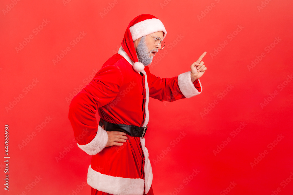 I told you! Side view of elderly man with gray beard wearing santa claus costume standing with attention sign, with finger warning gesture. Indoor studio shot isolated on red background.