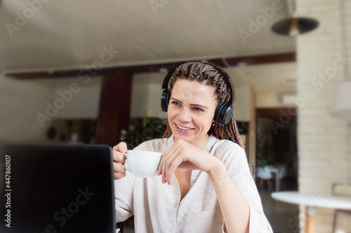 Young woman positively passionate about technology in wireless headphones sits at a table in a cozy cafe and uses modern devices.
