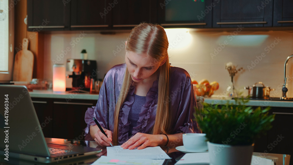 Blonde business woman in bathrobe signing documents in the kitchen at home. She also working on the computer.