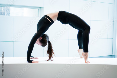 Young athletic flexible fitness woman practice chakrasana wheel, wearing black sportswear, in panoramic light window at home studio indoor on white background photo