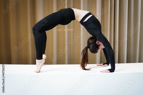 Young athletic flexible fitness woman practice chakrasana wheel, wearing black sportswear, in panoramic light window at home studio indoor on white background photo