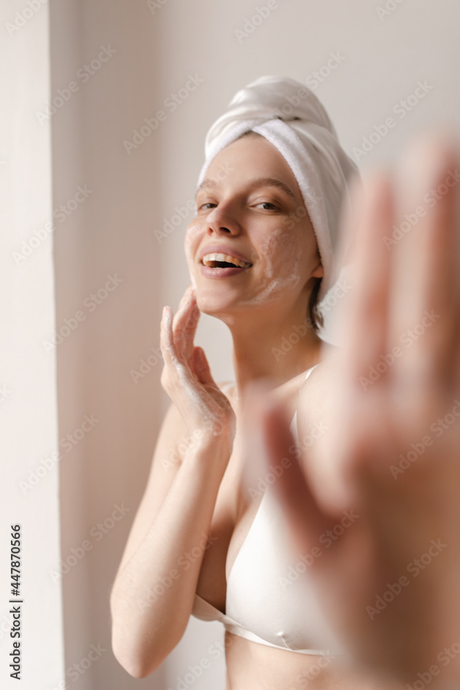 Lifestyle portrait of woman washing with one hand on foreground. Young lady in white bra smiling and looking straight forward while making body care procedures against light background Lifestyle portrait of woman washing with one hand on foreground. Young lady in white bra smiling and looking straight forward while making body care procedures against light background