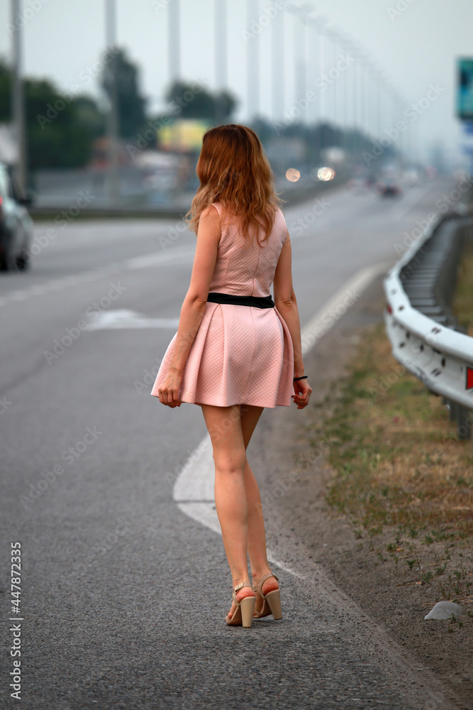 Slender woman in pink dress walking by the road from back