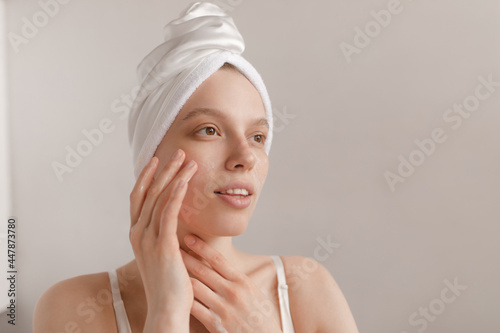 Gorgeous lady applying cream on pure skin. Girl with white towel wrapped hair, making procedures and looking aside isolated over light background. Cosmetology and skincare concept
