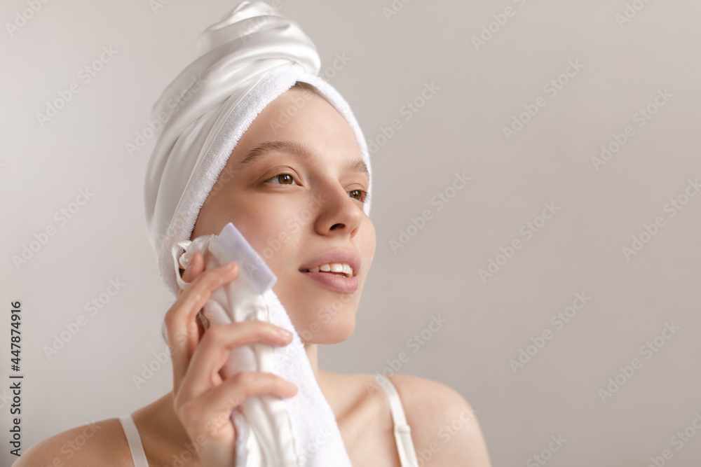 Cleansing and skincare concept. Lifestyle portrait of young lady making treatment procedures, wiping face with white towel while hair wrapped Cleansing and skincare concept. Lifestyle portrait of young lady making treatment procedures, wiping face with white towel while hair wrapped