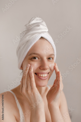 Pretty pale girl making procedures. Lady with white towel wrapped hair smiling and applying cream on pure face and looking into camera against light background