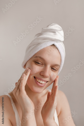 Young lady making treatment procedures. Smiling pretty girl applying lotion on face with hands and wearing towel, looking aside and posing against light background