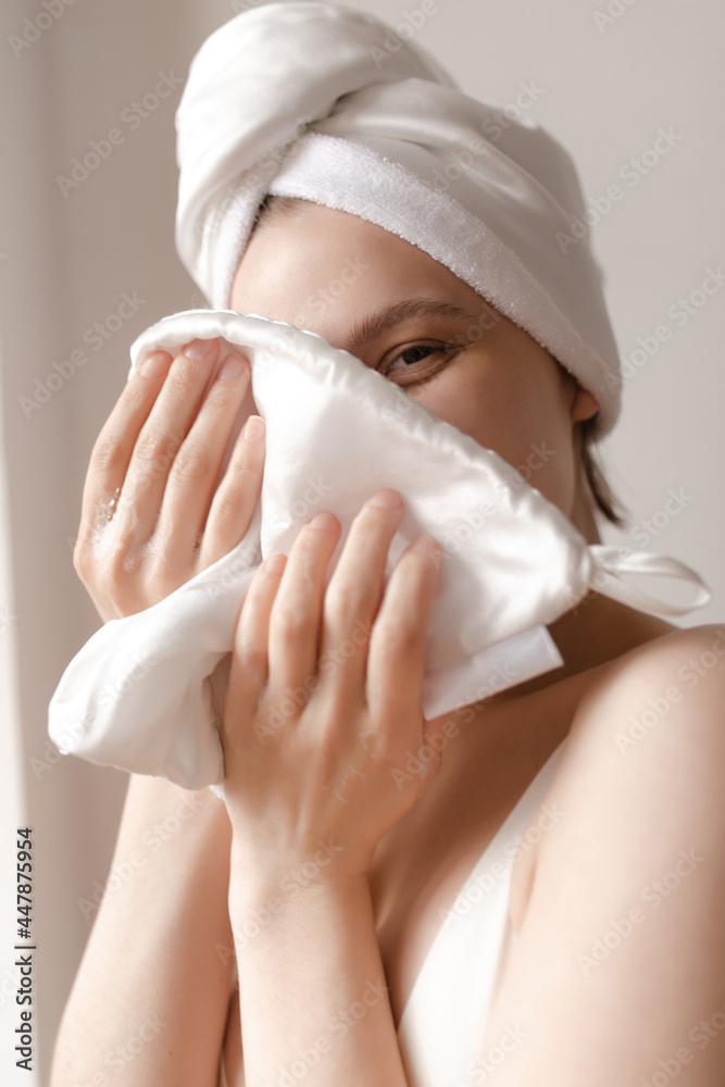 Skincare and wellness concept. Charming young lady with towel wrapped hair wiping face and looking into camera against light background Skincare and wellness concept. Charming young lady with towel wrapped hair wiping face and looking into camera against light background