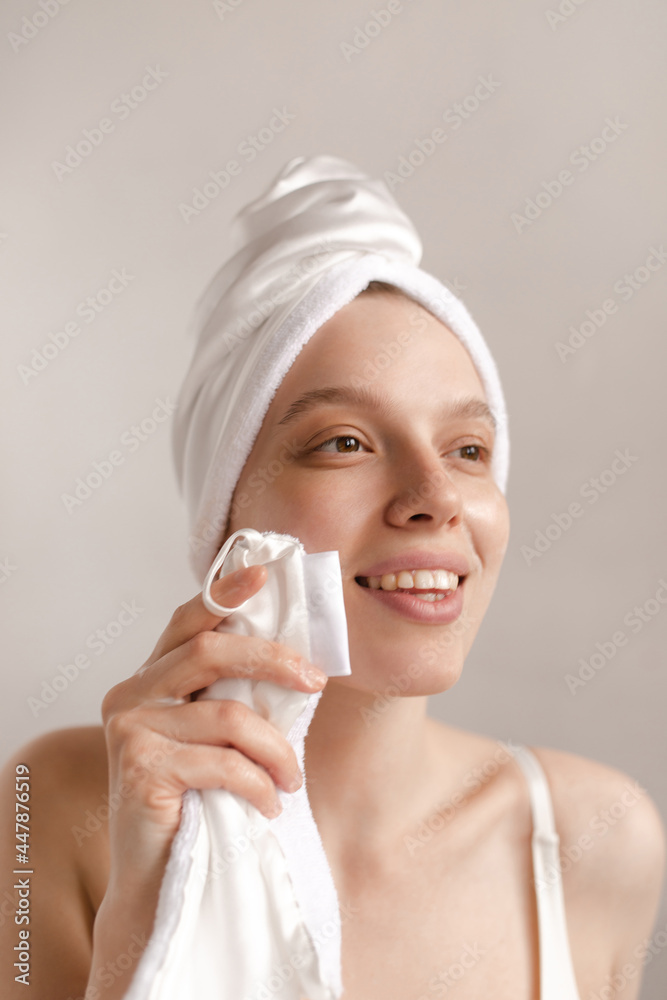 Pretty young girl wiping face. Charming lady with towel wrapped hair smiling, making cleansing procedures and looking aside against light background Pretty young girl wiping face. Charming lady with towel wrapped hair smiling, making cleansing procedures and looking aside against light background