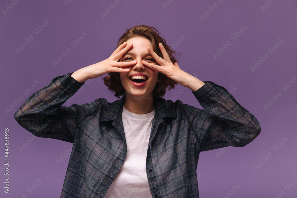 Fashion portrait of young girl isolated over violet background. Trendy short-haired lady with curls, white top and dark plaid shirt smiling and having fun while touching face
