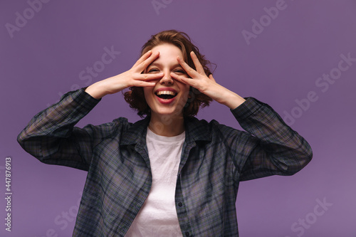 Fashion portrait of young girl isolated over violet background. Trendy short-haired lady with curls, white top and dark plaid shirt smiling and having fun while touching face