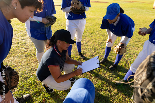 Diverse group of female baseball players in huddle around squatting female coach with clipboard photo