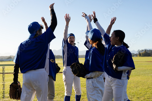 Diverse group of happy female baseball players raising hands on sunny baseball field before game photo