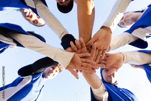 Diverse group of happy female baseball players stacking hands on sunny baseball field before game