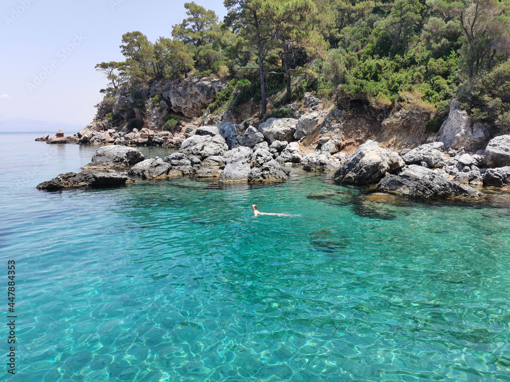 Young girl in bikini swims on the azure beach with rocks and pine trees. The Aegean sea. Turkey, Kusadasi.