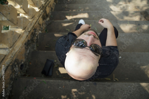 Young handsome man dressed casually sitting on the stairs in a park. Overhead shot looking down on bald cool guy in sunglasses relaxing outdoors