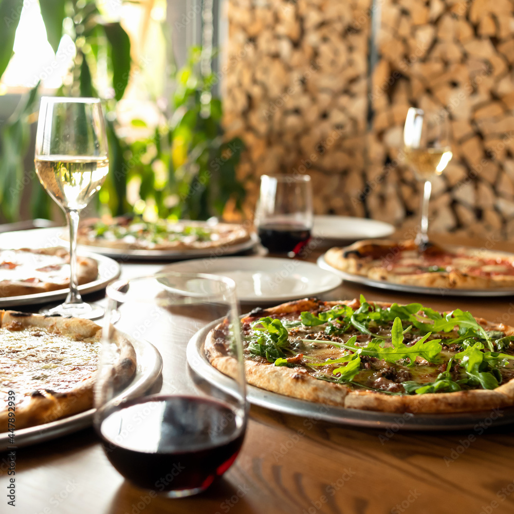 Pizza party. Assorted five different pizzas on wooden restaurant table with glasses of wine on it. Woodpile of firewood on blurred background. Side view.