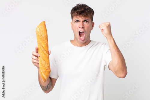 young handsome man shouting aggressively with an angry expression and holding a bread baguette